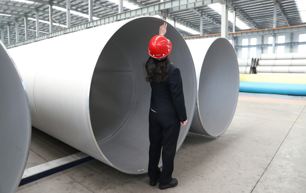 Engineer in a hard hat inspecting large steel pipe sections in a factory