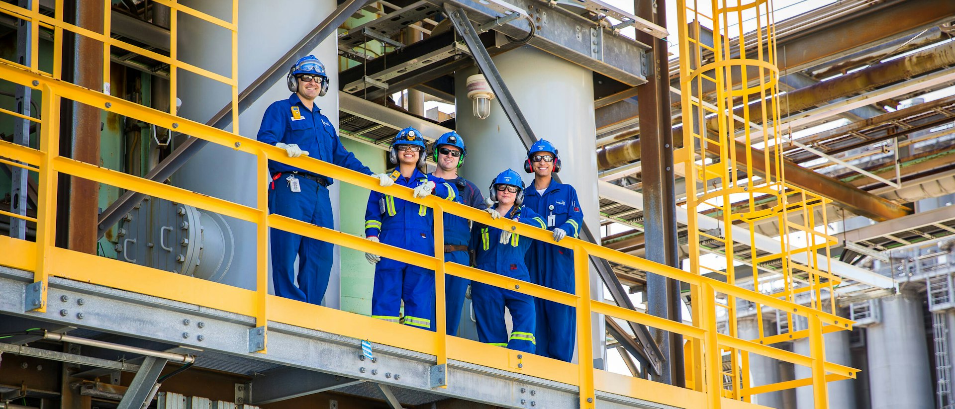 Engineering team in coveralls, hard hats and safety glasses on a platform at an industrial plant